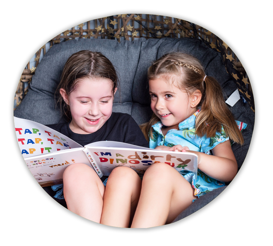 Two children sitting on a chair smiling as they read a book together.