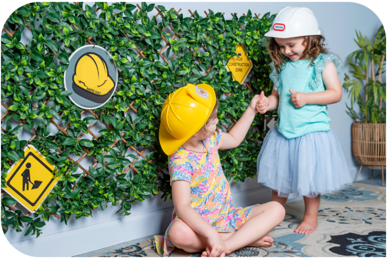 Two children in hard hats play in front of a wall covered in vines and safety signs.