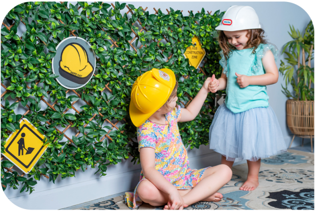 Two children in hard hats play in front of a wall covered in vines and safety signs.